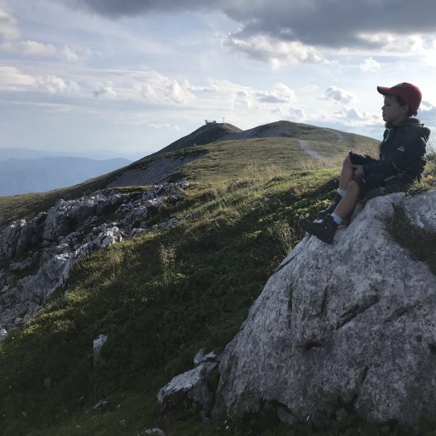 Ein Junge mit roter Kappe sitzt auf einem Felsen und blickt über eine grüne Berglandschaft.