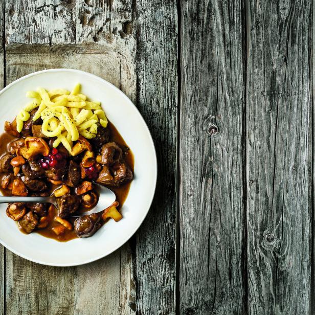 Ein Teller mit Wildgulasch, Spätzle und Preiselbeeren auf einem Holztisch.