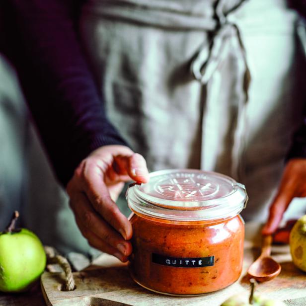 Eine Person mit Schürze hantiert mit einem Glas Quittenmarmelade auf einem Holztisch.
