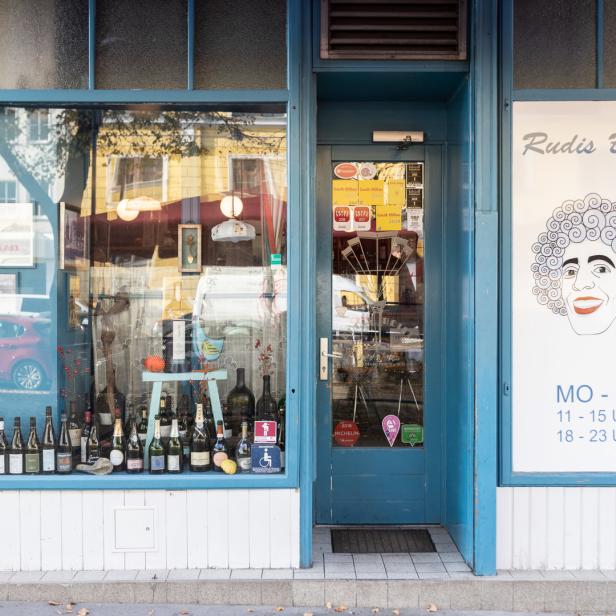 Die Fassade von „Rudis Beisl“ mit Weinflaschen im Schaufenster und Öffnungszeiten an der Tür.