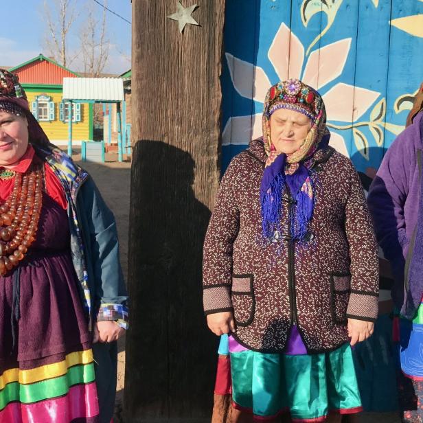 Drei Frauen in traditioneller burjatischer Kleidung stehen vor Holzhäusern.
