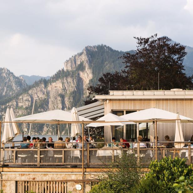 Gäste sitzen auf der Terrasse eines Restaurants am See mit Blick auf die Berge.