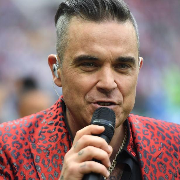 Robbie Williams singt in einem roten Leopardenmuster-Sakko in ein Mikrofon.