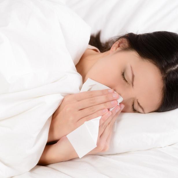 Eine Frau liegt mit einer Erkältung im Bett und putzt sich die Nase.