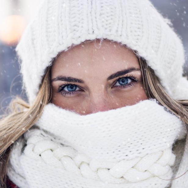 Eine Frau mit blauen Augen trägt eine weiße Mütze und einen Schal im Schnee.