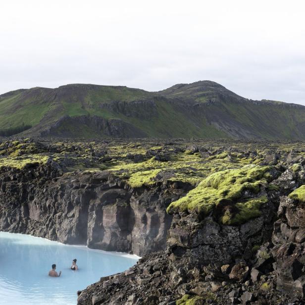 Zwei Personen baden in einem türkisfarbenen Becken, umgeben von Lavafelsen und grünen Hügeln.