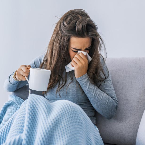 Eine erkältete Frau sitzt mit einer Tasse Tee und einem Taschentuch auf dem Sofa.