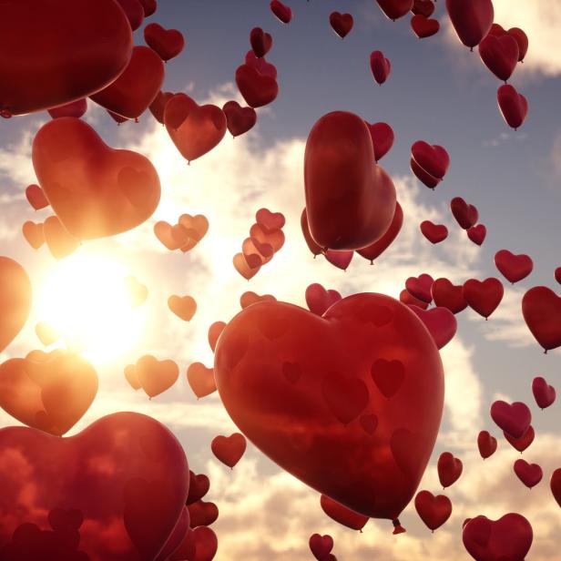 Viele rote Herzballons steigen vor einem bewölkten Himmel auf.
