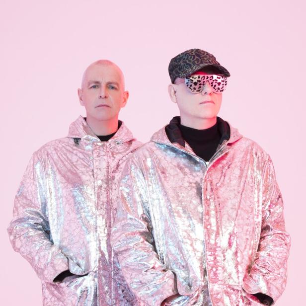 Die Pet Shop Boys in glänzenden Jacken vor einem rosa Hintergrund.