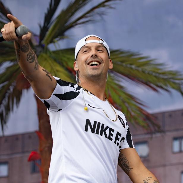 Kontra K tritt in einem Nike-Trikot mit Tätowierungen und einer Kappe auf einer Bühne auf.