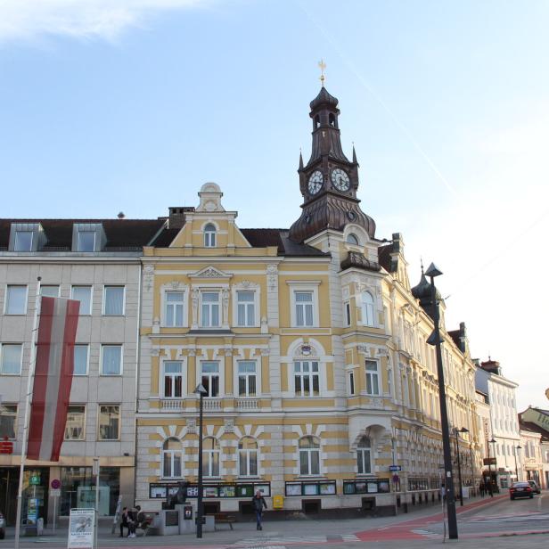 Historisches gelbes Gebäude mit Uhrturm und mehreren Flaggen davor an einem Platz bei Tageslicht.