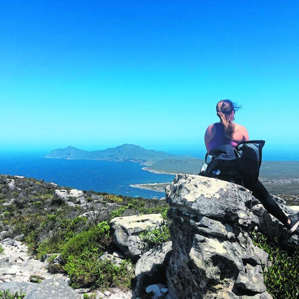 Eine Frau sitzt mit einem Rucksack auf einem Felsen und blickt auf das Meer.