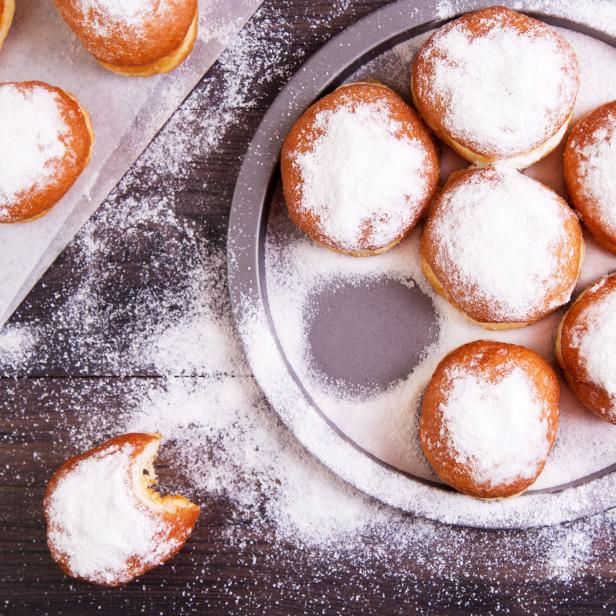 Mehrere mit Puderzucker bestäubte Krapfen auf einem Backblech und einem Küchentuch.