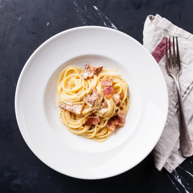 Ein Teller Spaghetti Carbonara mit Speckwürfeln und Pfeffer auf einem dunklen Tisch.