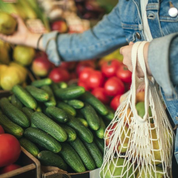Eine Person wählt auf einem Markt Birnen und Gurken aus und hält eine Stofftasche.