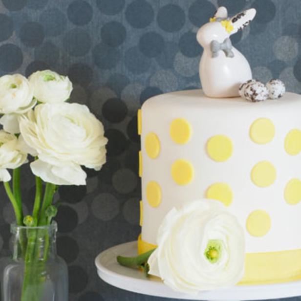 Ein dekorativer Kuchen mit gelben Punkten und einem Hasen, neben weißen Blumen in einer Vase.