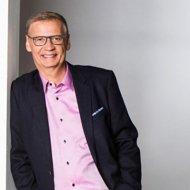 Günther Jauch lächelt in einem dunklen Sakko und einem hellrosa Hemd vor einem hellen Hintergrund.