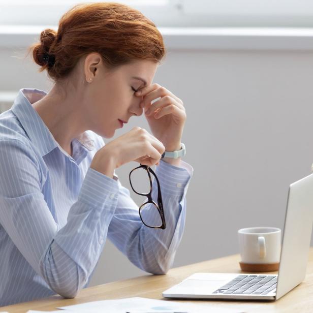 Eine Frau sitzt erschöpft vor ihrem Laptop und hält ihre Brille in der Hand.