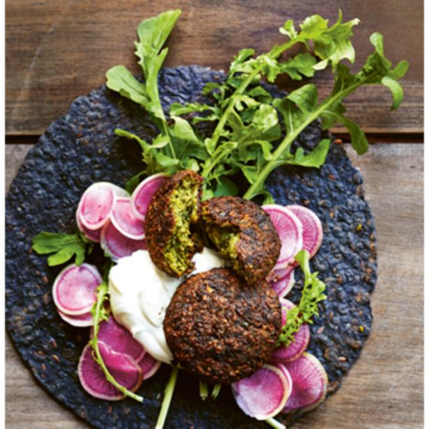 Drei Falafel liegen mit Radieschen, Salat und einer weißen Soße auf einem dunklen Teller.