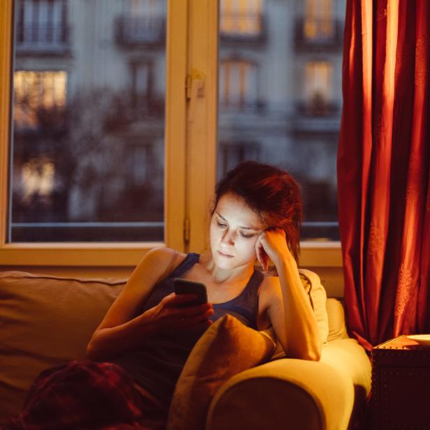 Eine Frau sitzt abends auf einem Sofa und schaut auf ihr Smartphone.