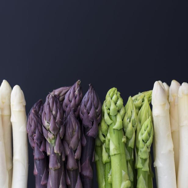 Verschiedene Sorten Spargel, darunter weißer, grüner und violetter Spargel, vor einem dunklen Hintergrund.