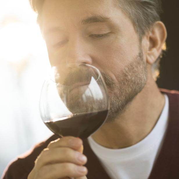Ein Mann mit Bart riecht an einem Glas Rotwein.