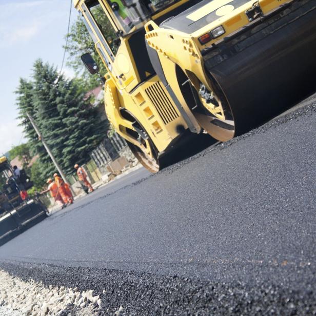 Gelber Straßenwalze verdichtet frisch aufgetragenen schwarzen Asphalt auf einer Straße, im Hintergrund Bagger und Bäume.