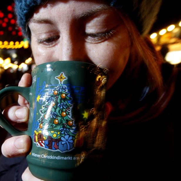 Eine Person trinkt aus einer grünen Tasse mit Weihnachtsbaum-Motiv auf einem festlich beleuchteten Weihnachtsmarkt.