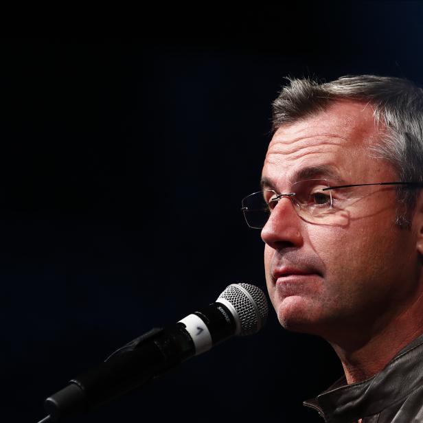 Ein Mann mit Brille und grauen Haaren spricht in ein Mikrofon, dunkler Hintergrund.