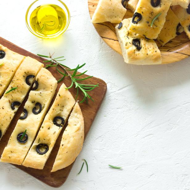Geschnittene Focaccia mit Oliven und Rosmarin auf einem Holzbrett, dazu Olivenöl.