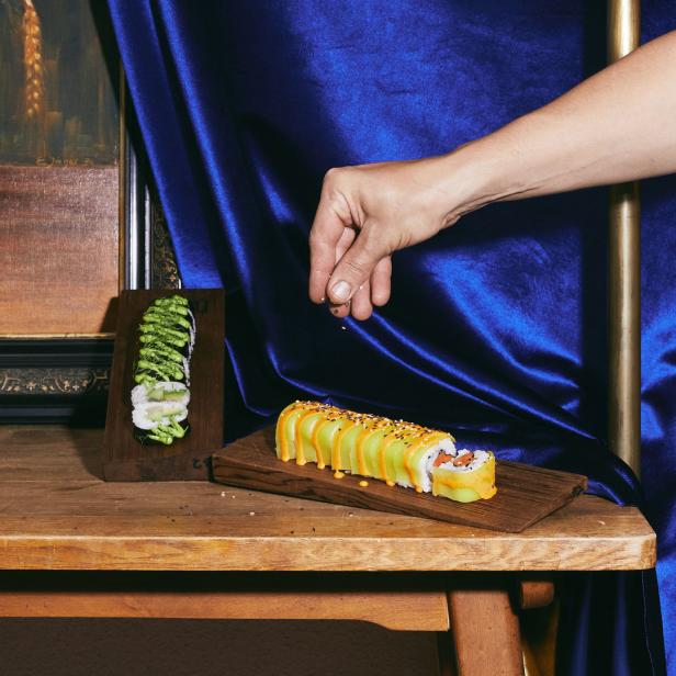 Eine Hand streut Sesam über eine Sushi-Rolle mit Avocado auf einem Holztisch.