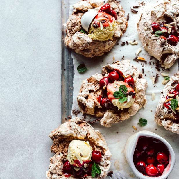 Mehrere Schokoladen-Pavlovas mit Kirschen, Eis und Minze auf einem Backblech.