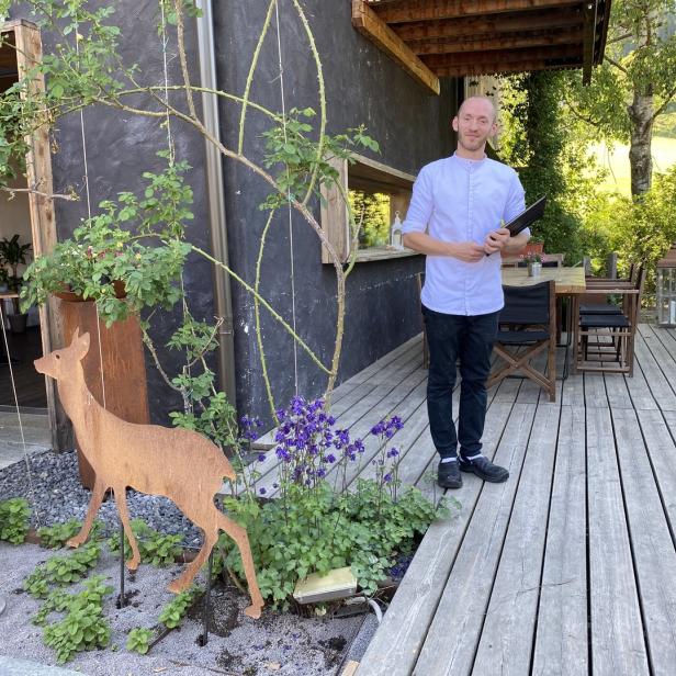 Ein Mann in Kochkleidung steht vor einem Restaurant mit Holzterrasse und Gartendekoration.