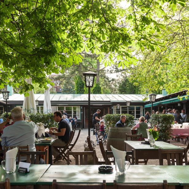 Ein Biergarten mit Gästen unter schattenspendenden Bäumen.