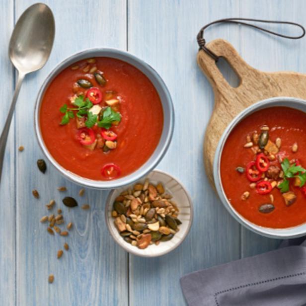Zwei Schalen mit Tomatensuppe, garniert mit Kräutern, Samen und Chili, auf einem Holztisch.