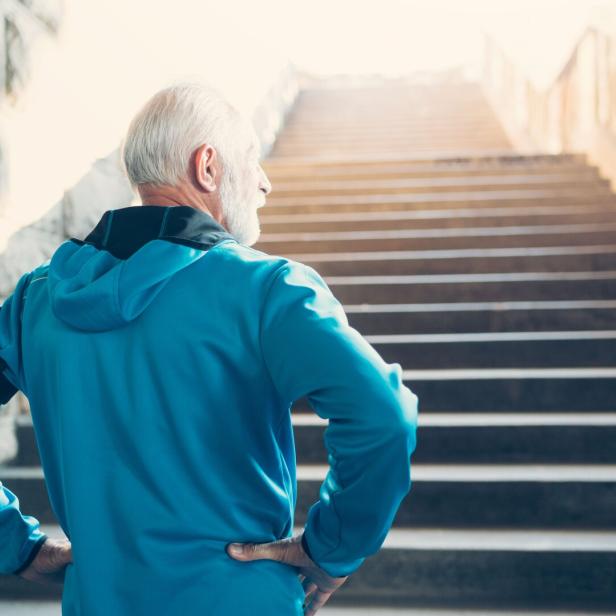 Ein älterer Mann in Sportkleidung steht mit den Händen in der Hüfte vor einer langen Treppe.
