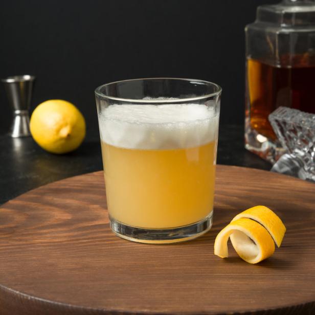 Ein Whiskey Sour Cocktail auf einem Holztablett mit einer Zitronenspirale.