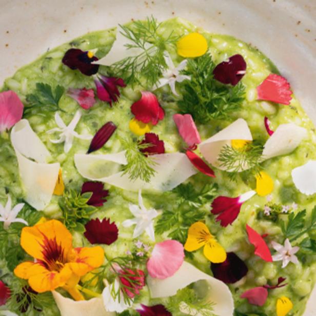 Ein grünes Risotto ist mit essbaren Blüten und Kräutern auf einem Teller angerichtet.