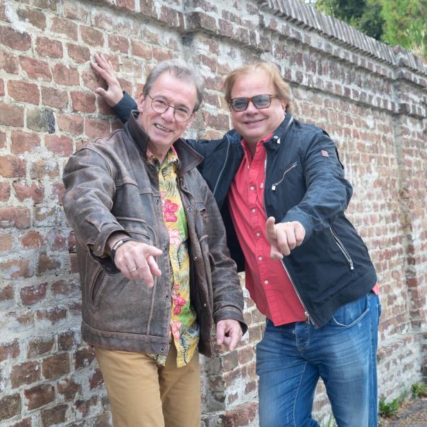 Zwei Männer posieren lächelnd vor einer Backsteinmauer.