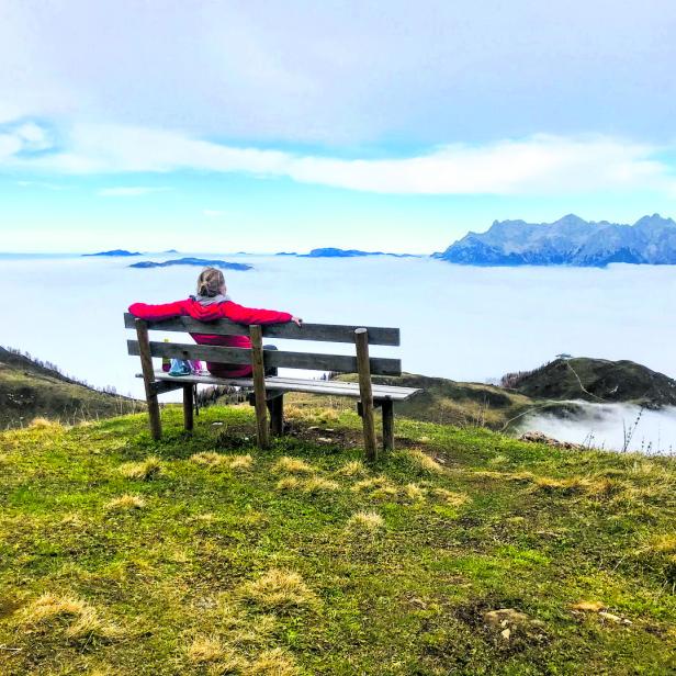 Eine Frau sitzt auf einer Bank und blickt auf eine Nebellandschaft mit Bergen.