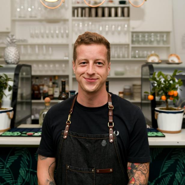 Ein lächelnder Barkeeper steht vor einer Bar mit Getränken und Dekorationen.