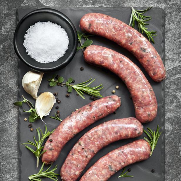 Rohe Bratwürste liegen mit Salz, Knoblauch und Kräutern auf einem dunklen Teller.