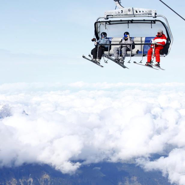 Drei Skifahrer sitzen in einem Sessellift über einer Wolkendecke.
