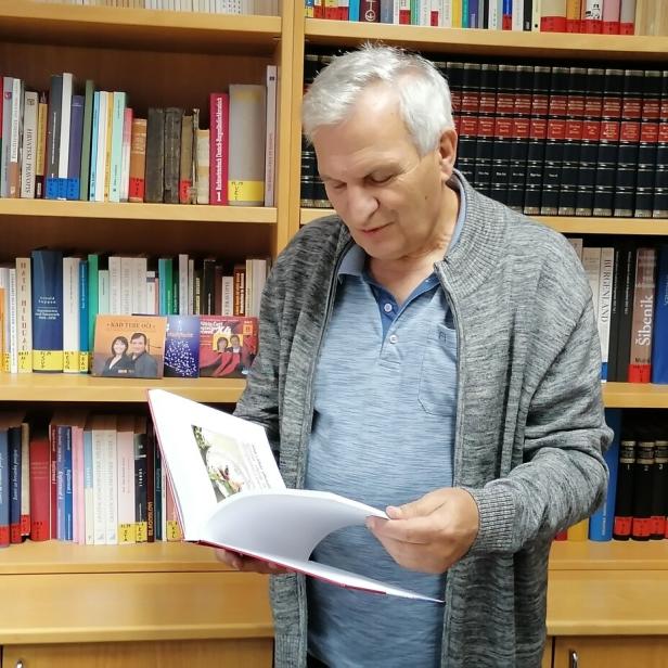 Ein älterer Mann steht vor einem Bücherregal und liest in einem aufgeschlagenen Buch.