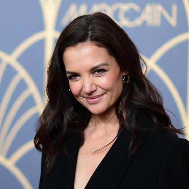 Katie Holmes lächelt vor einem goldenen Art-Déco-Hintergrund.