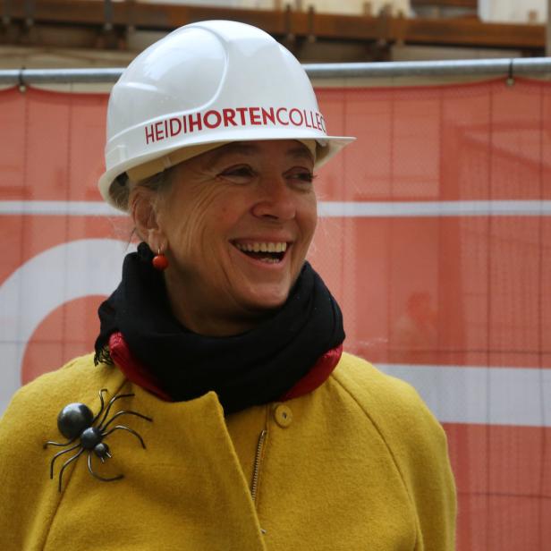 Eine lächelnde Frau mit Bauhelm und gelbem Mantel vor einem Bauzaun.
