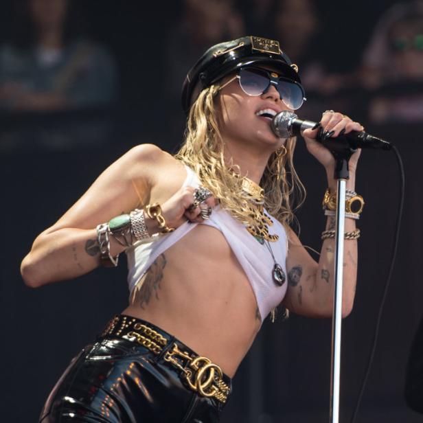 Miley Cyrus tritt in glänzender Hose, bauchfreiem Top und Mütze auf einer Bühne auf.