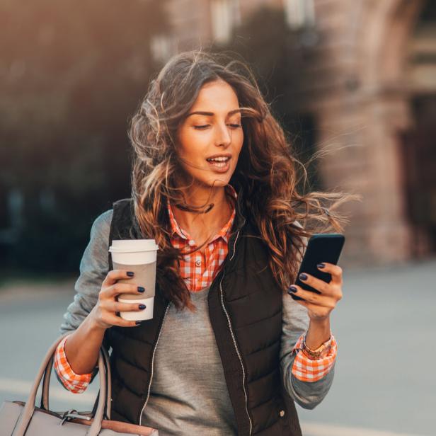 Eine Frau geht mit einem Kaffee und einem Handy in der Hand die Straße entlang.