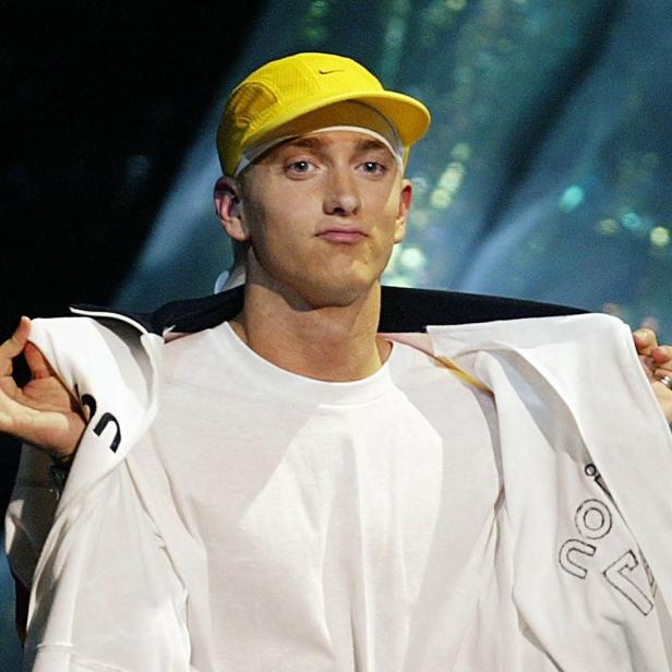 Eminem mit gelber Kappe und weißer Jacke auf einer Bühne.