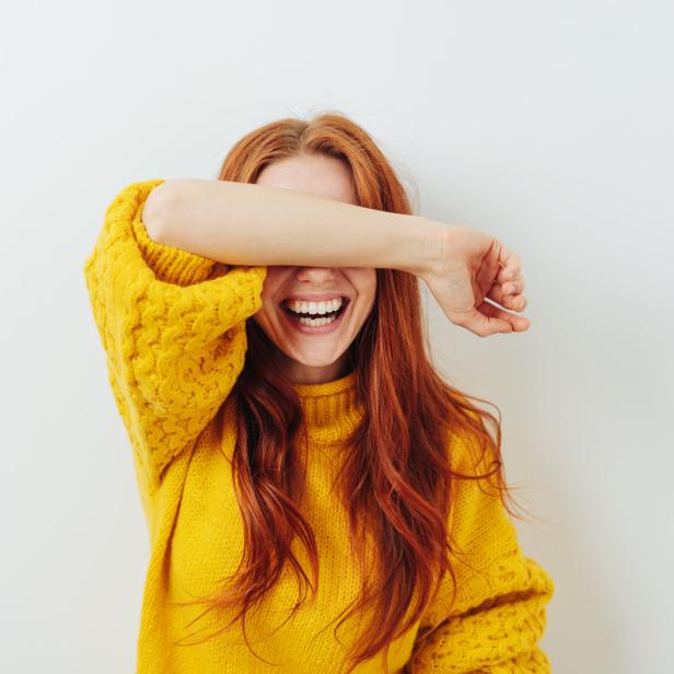 Eine lachende Frau mit roten Haaren und gelbem Pullover verdeckt ihr Gesicht mit dem Arm.